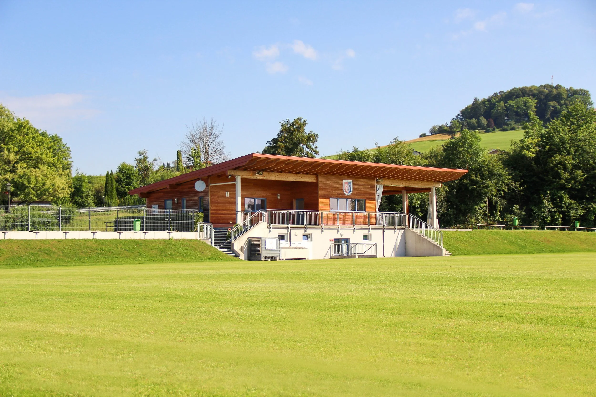 Ein modernes, holzverkleidetes Verwaltungsgebäude auf einem Sportplatz, umgeben von grüner Landschaft und Hügeln im Hintergrund.