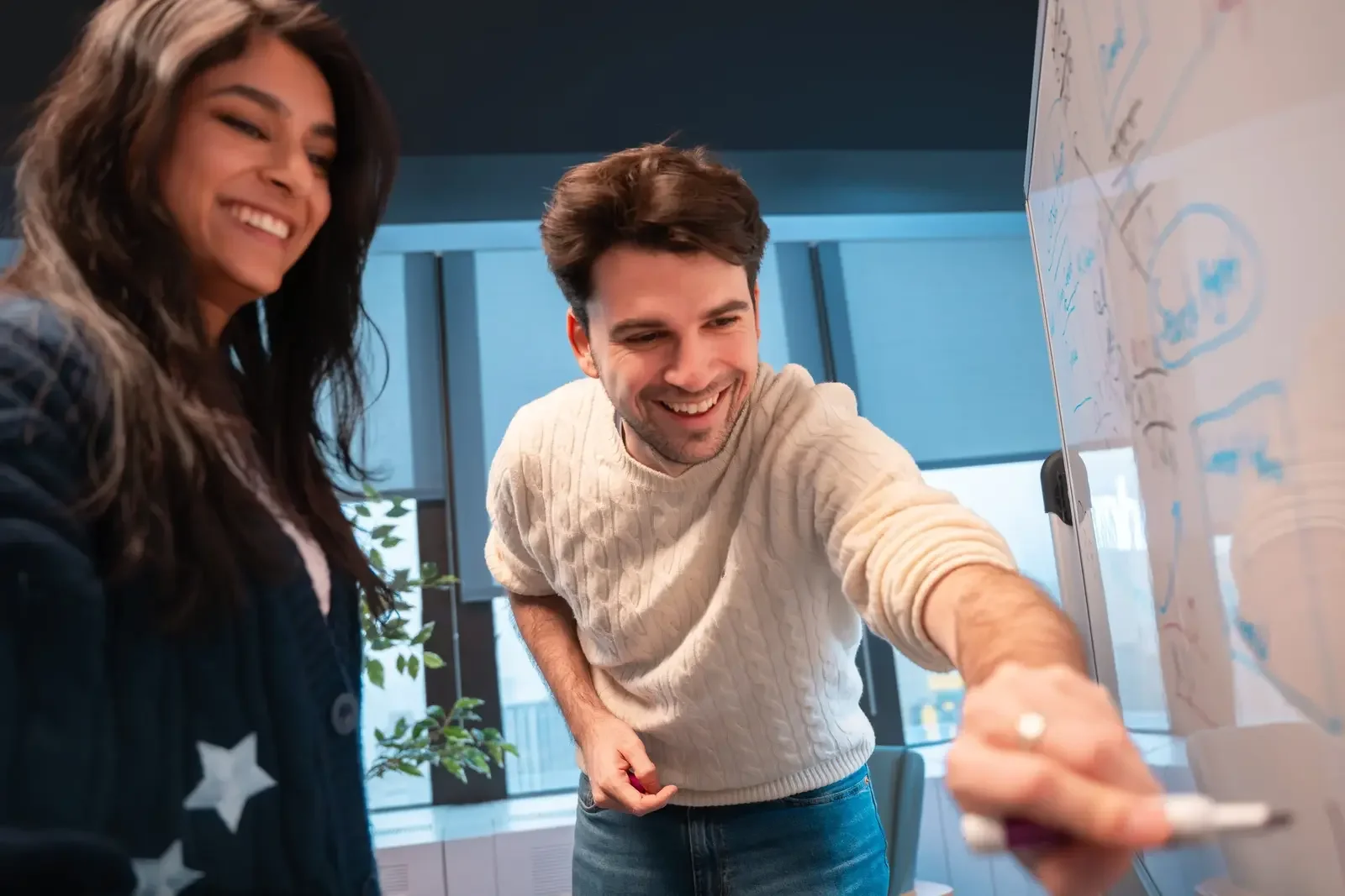 Two people, a woman and a man, smiling as they look at and point to a whiteboard with drawings in an office setting.