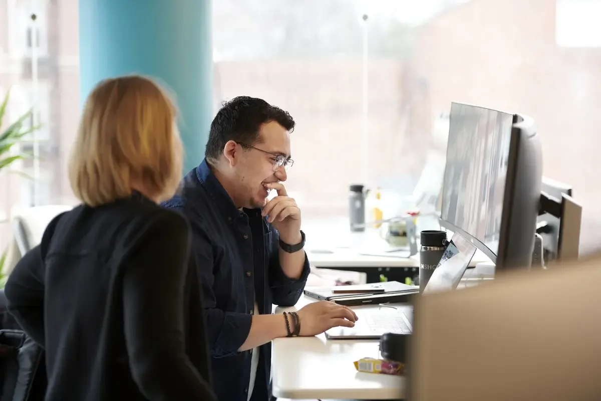 Two coworkers collaborate at a desk in an office, looking at a computer monitor and smiling.