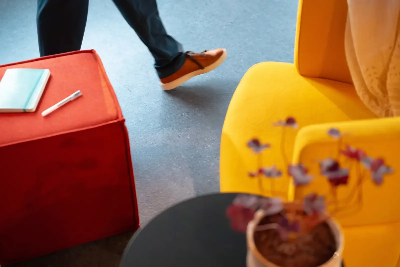 Part of a person's legs with black pants and brown shoes walking past a red box with a notebook and pen on top, yellow chairs, and a table with small flower arrangements.