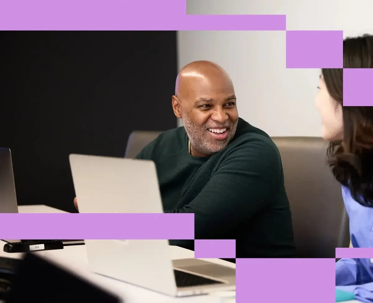 A man and a woman sitting at a table, talking and smiling in a business meeting, with laptops in front of them.