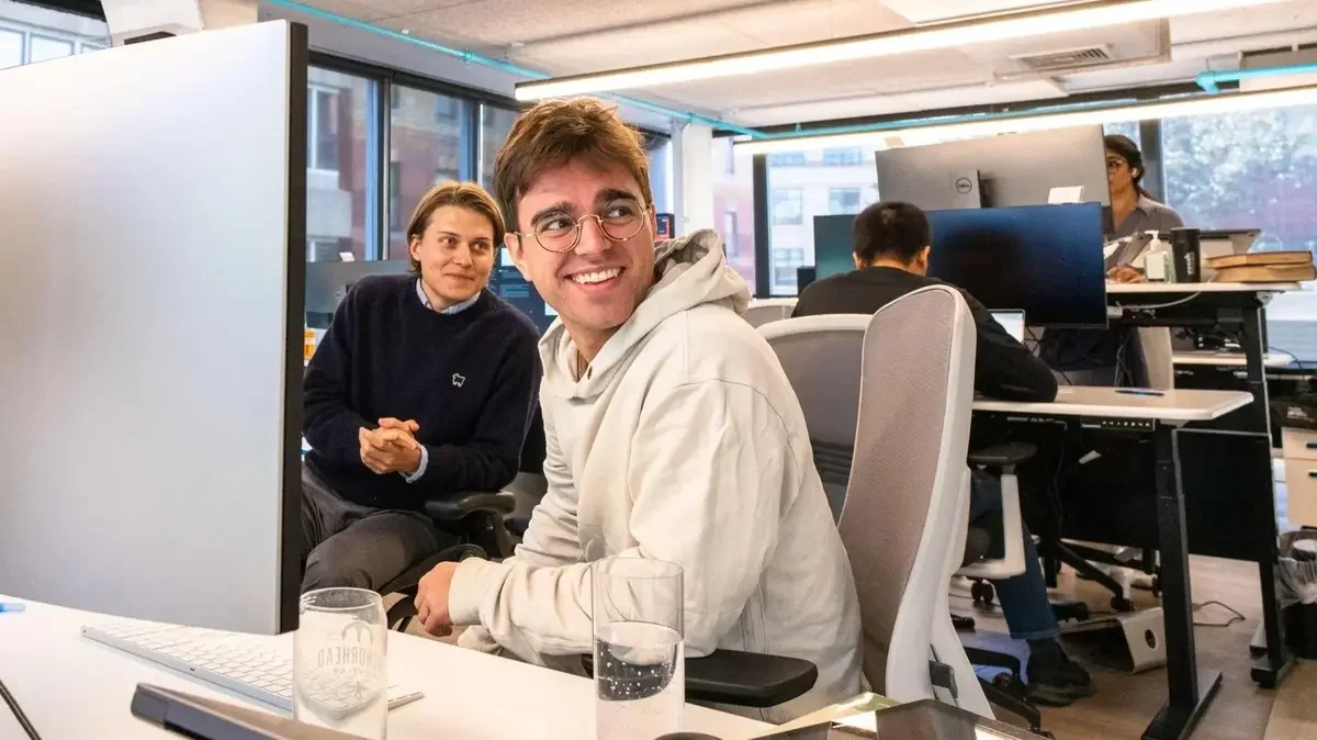 Office workers sitting at desks in a modern workspace with large windows, two people smiling at each other, and others working on computers.