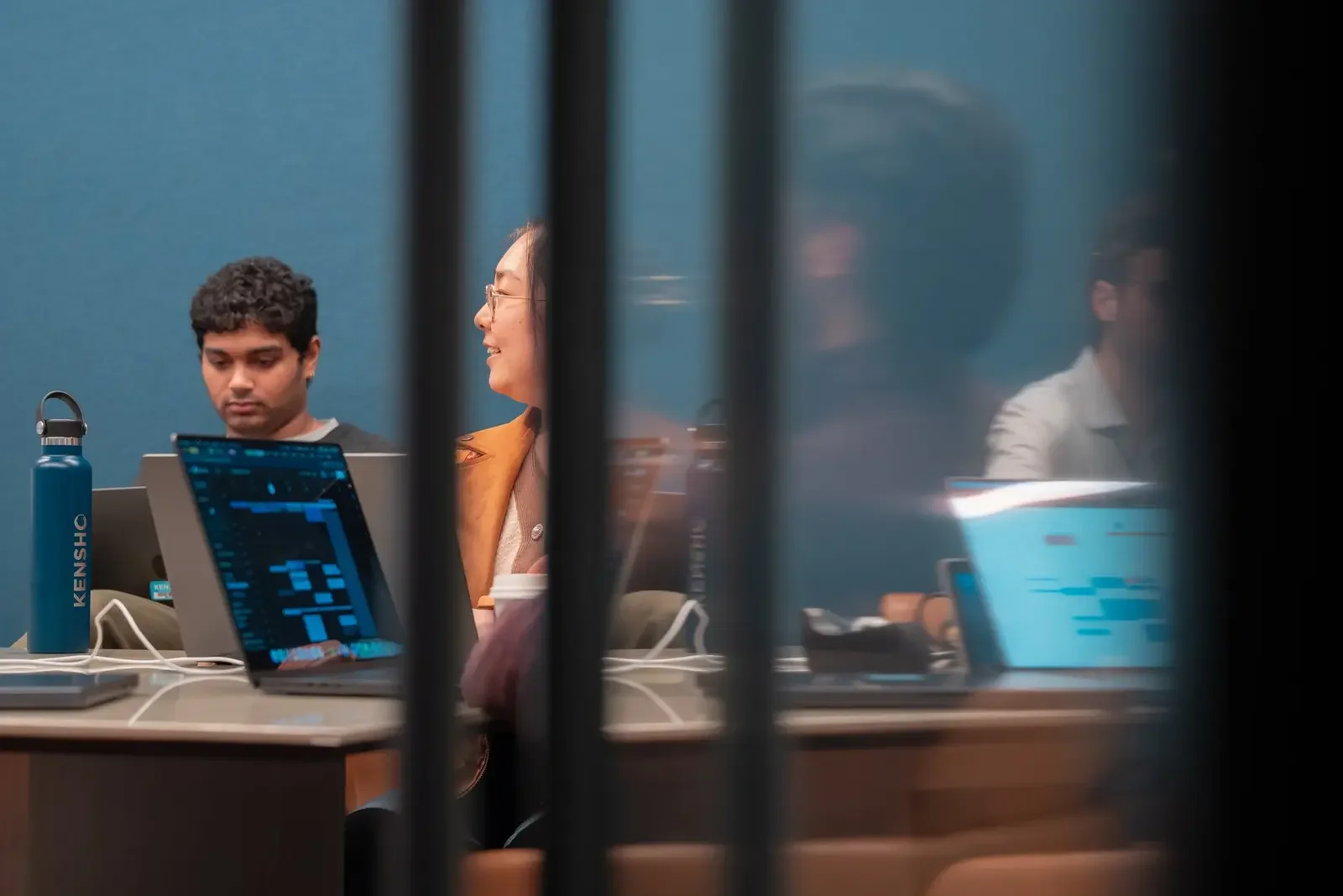 View through a partially obscured window showing three people sitting at a table with laptops and drinks in a meeting or workspace setting.