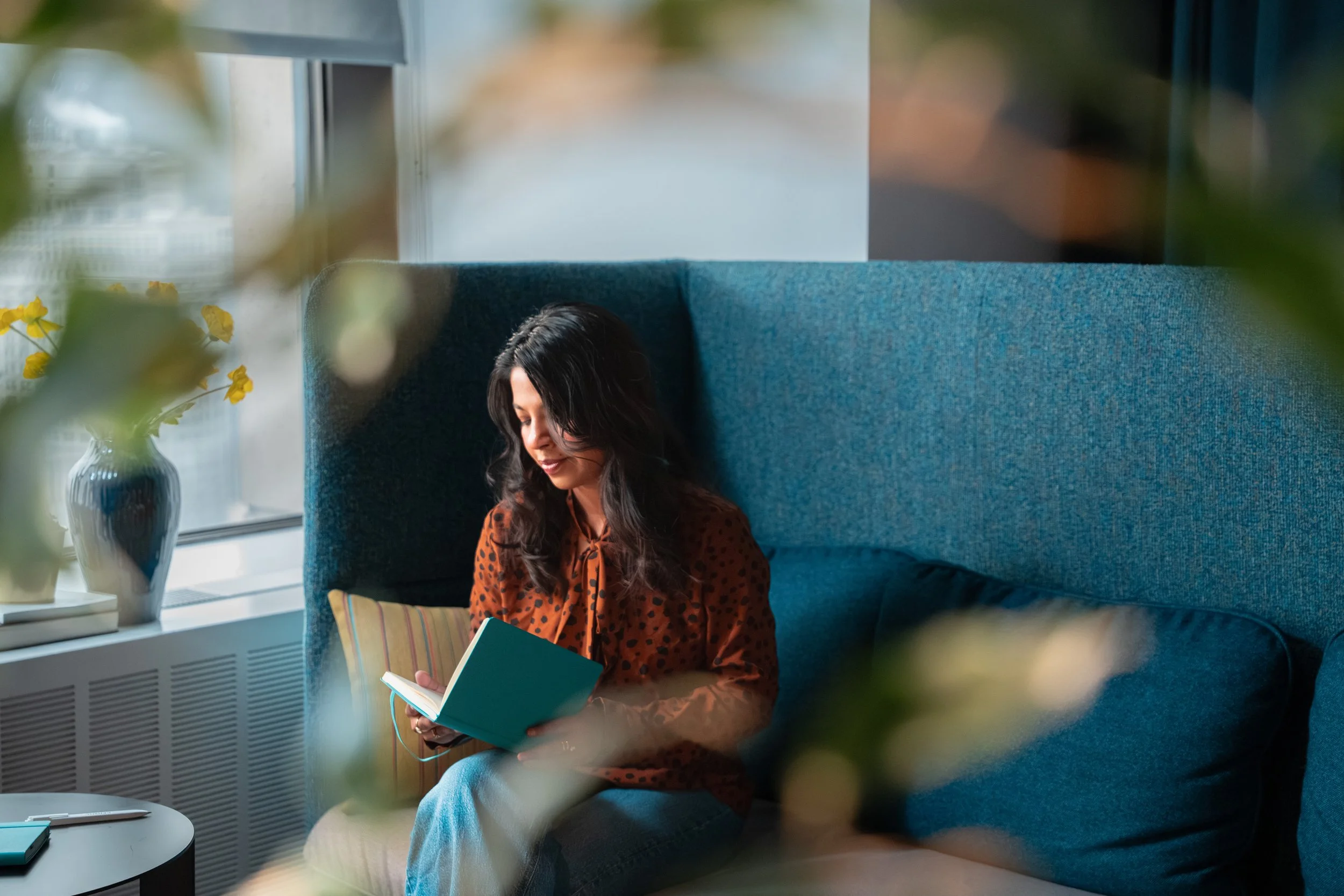 A woman with dark, wavy hair, wearing a rust-colored leopard print blouse, sits on a teal sofa reading a book. She is smiling with her eyes closed. The scene is bright with natural light from the large window beside her, which shows a vase with yellow flowers on the windowsill. Blurred greenery and leaves are in the foreground.