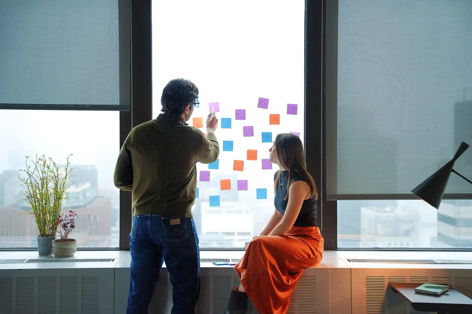 A man and woman discussing colored sticky notes on a glass window in a high-rise office. The woman is sitting on a windowsill, and the man is standing and pointing at the notes.