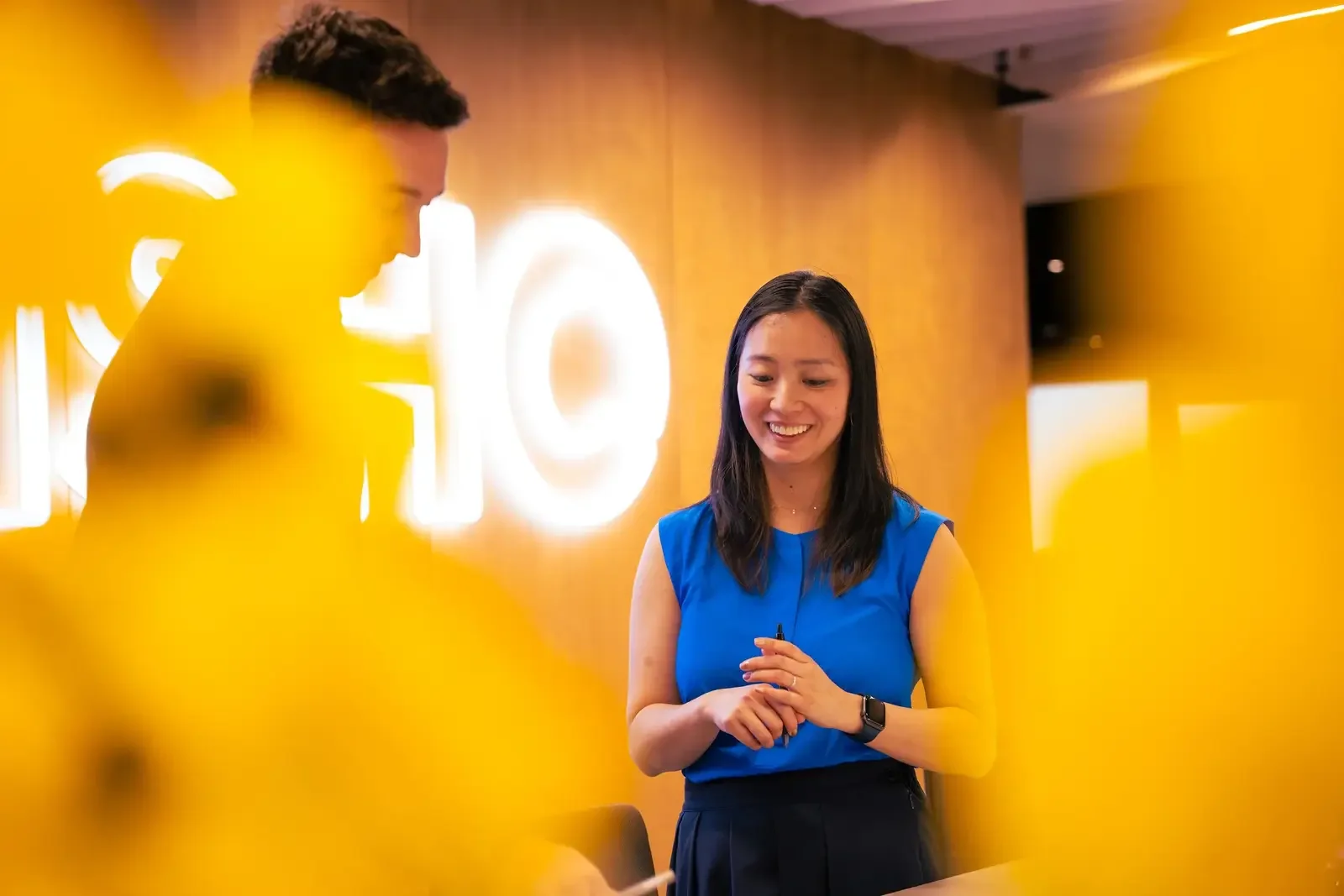 A woman in a blue sleeveless shirt is smiling and talking to a man in a yellow shirt at what appears to be a professional setting with a bright yellow wall and illuminated signage in the background.