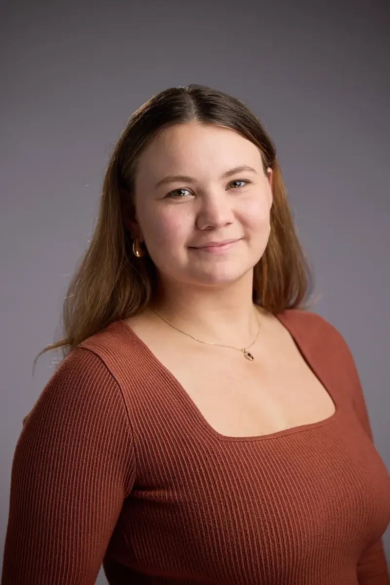 A young woman with light brown hair, wearing a rust-colored ribbed top, gold hoop earrings, a delicate gold necklace, and a subtle smile, posing against a neutral gray background.
