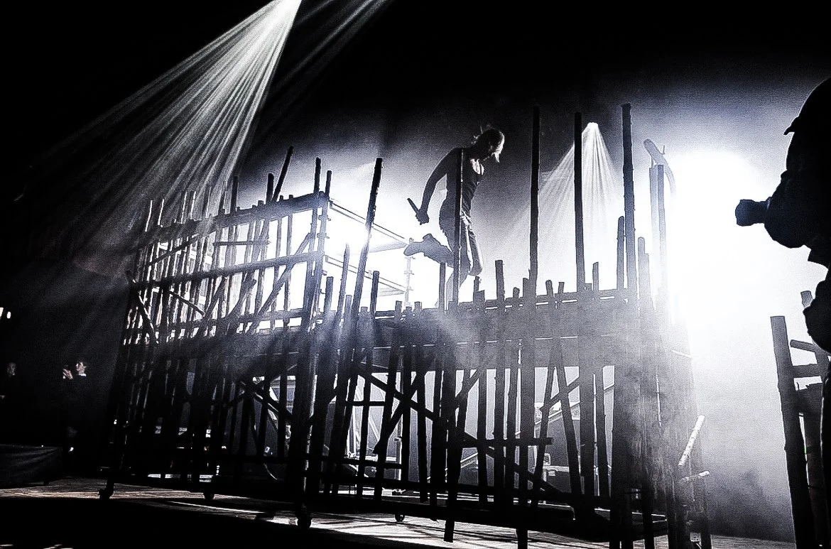 A construction site with a worker walking on scaffolding, silhouetted against bright light. The scene is dark with beams of light shining down.