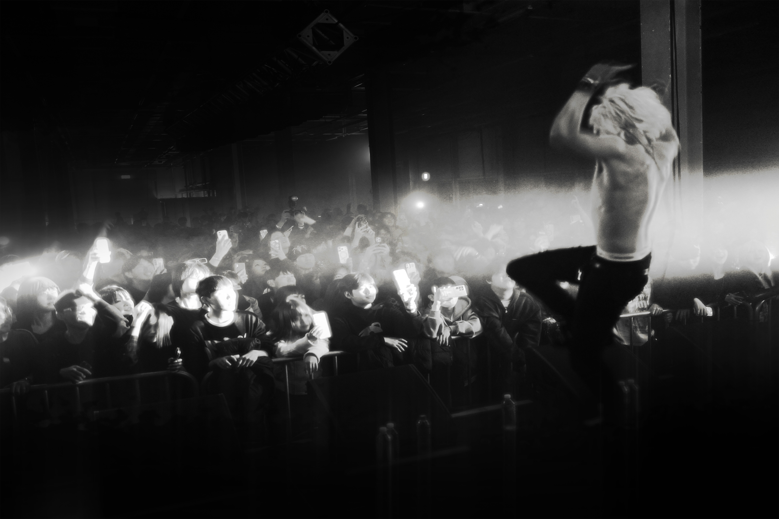 A male performer with long hair, shirtless, standing on a stage with one foot on a raised platform, facing away from the camera. The crowd in front of him is watching and taking photos with their phones, illuminated by stage lights.