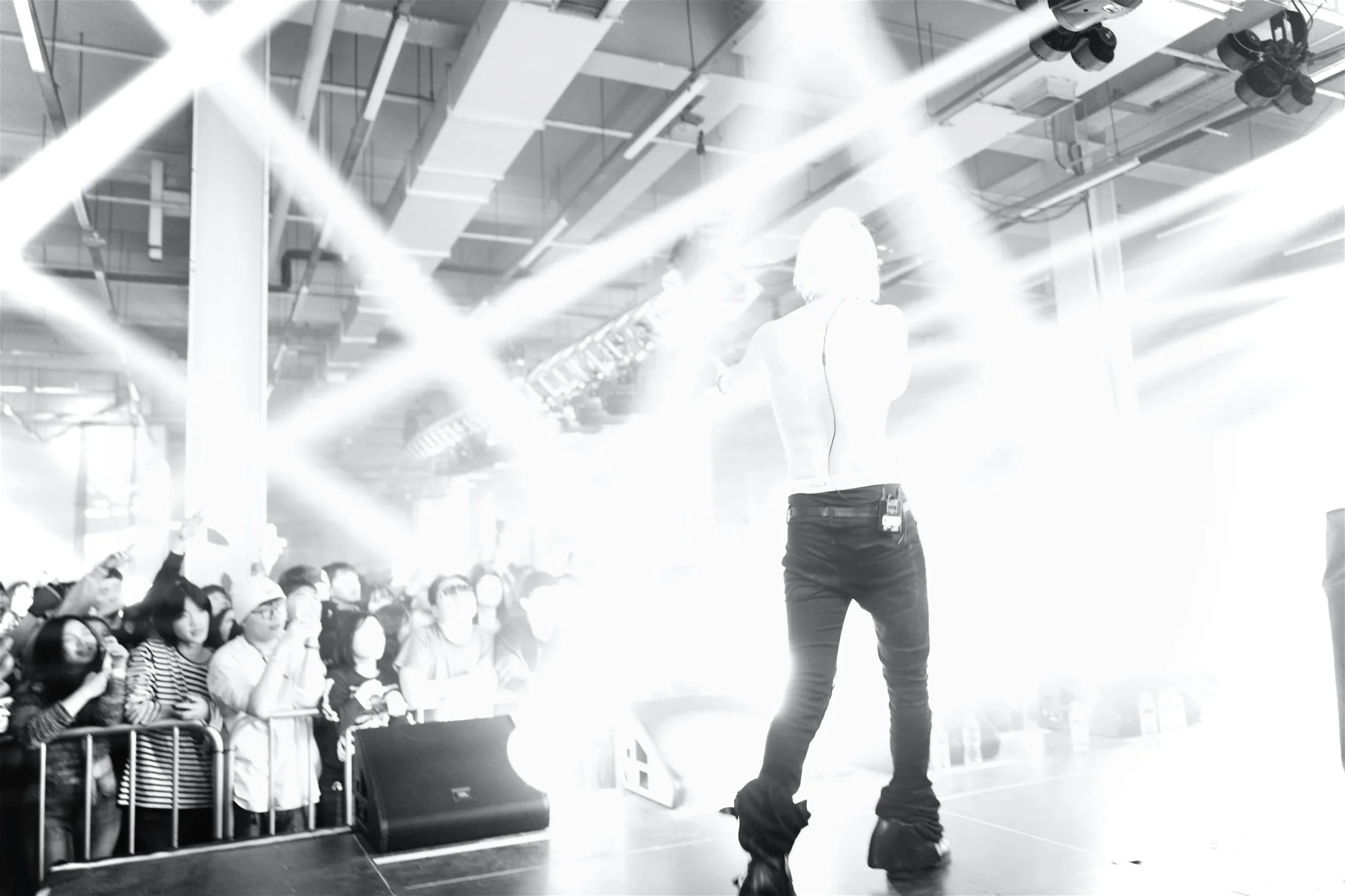 Performer on stage with bright lights shining, facing an audience behind a barricade, in a concert or event setting.