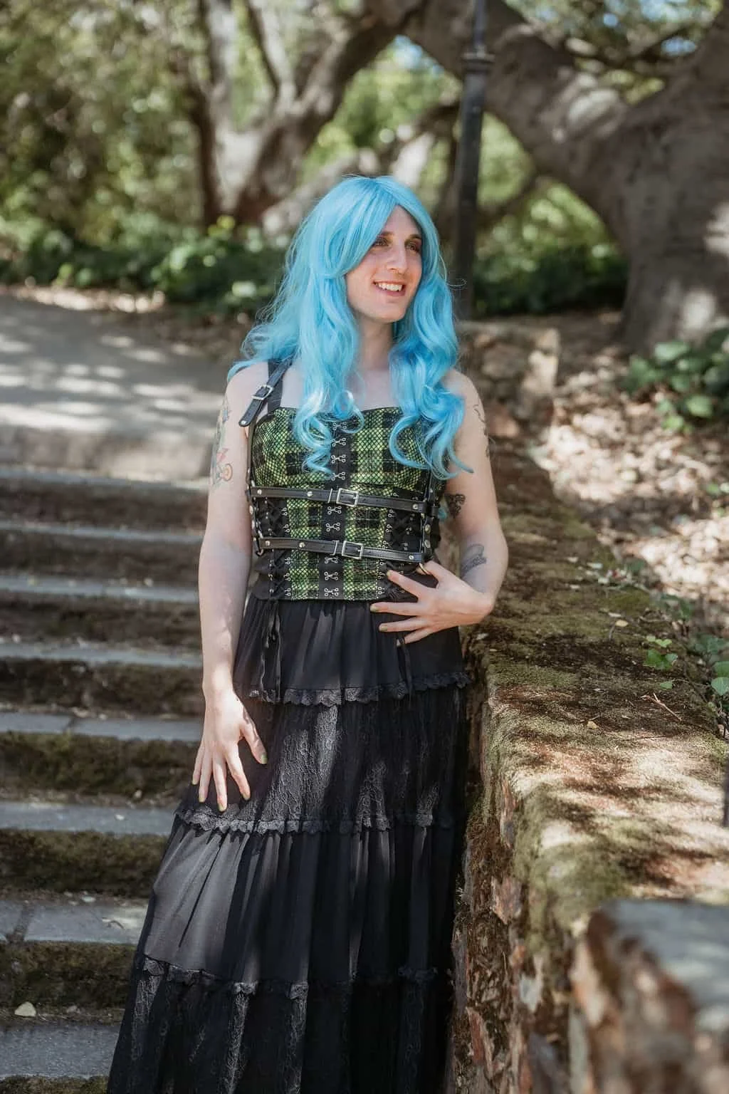 Calla wearing a bright blue wig wearing a dark green plaid corset as she's standing on mossy stairs.