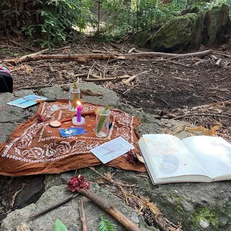 A Reiki ritual spread in a Redwood forest with a purple candle, clipper card, tarot cards, and drawings in a notebook