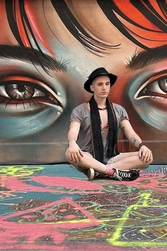 A queer medium siting cross legged, gazing confidently into the camera, posed in-front of graffiti art of a woman’s face