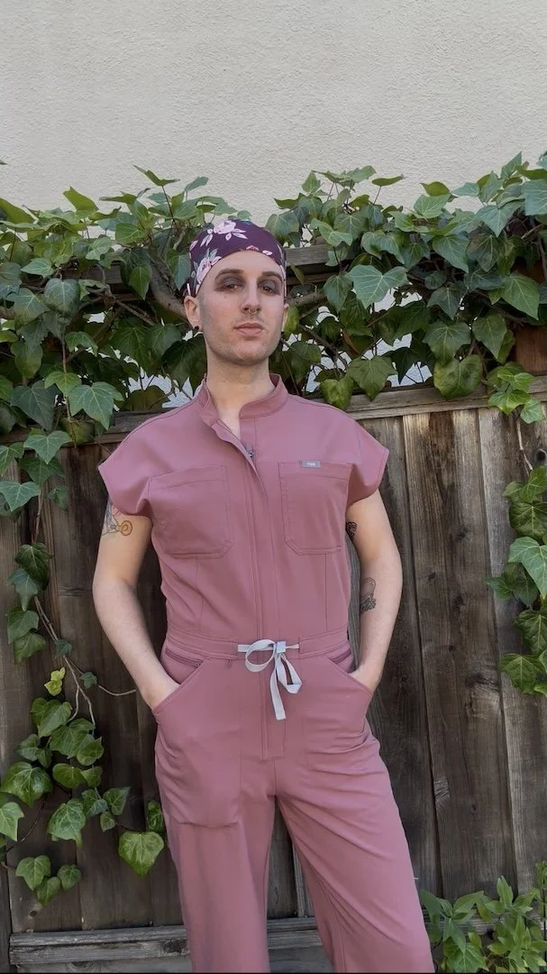 Transwoman wearing a pink Figs scrub jumpsuit and scrub cap posing powerfully in front of a fence covered in ivy