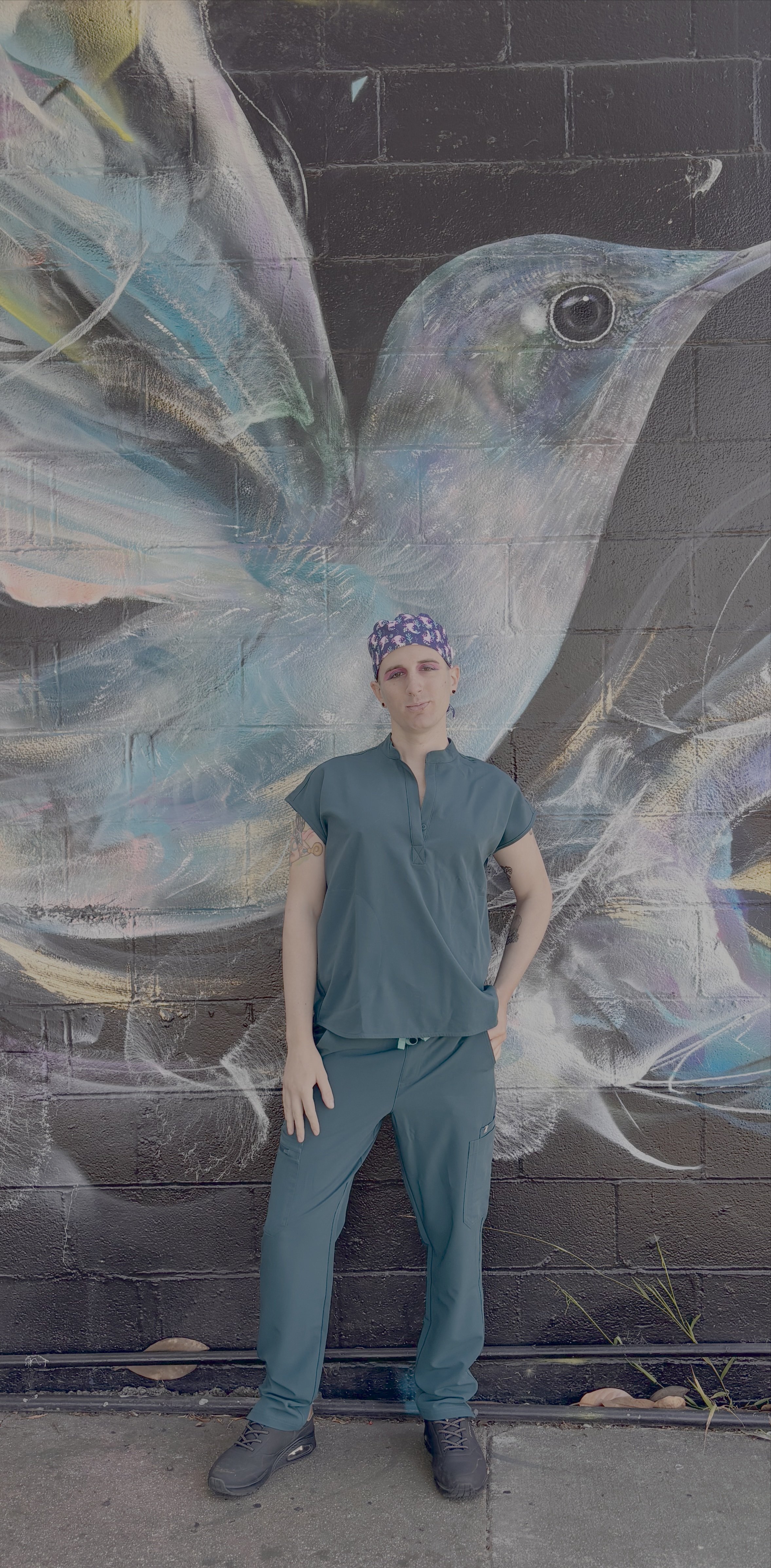 Transwoman dressed in Caribbean blue Figs scrubs posing in front of a painting of a hummingbird on the side of a building