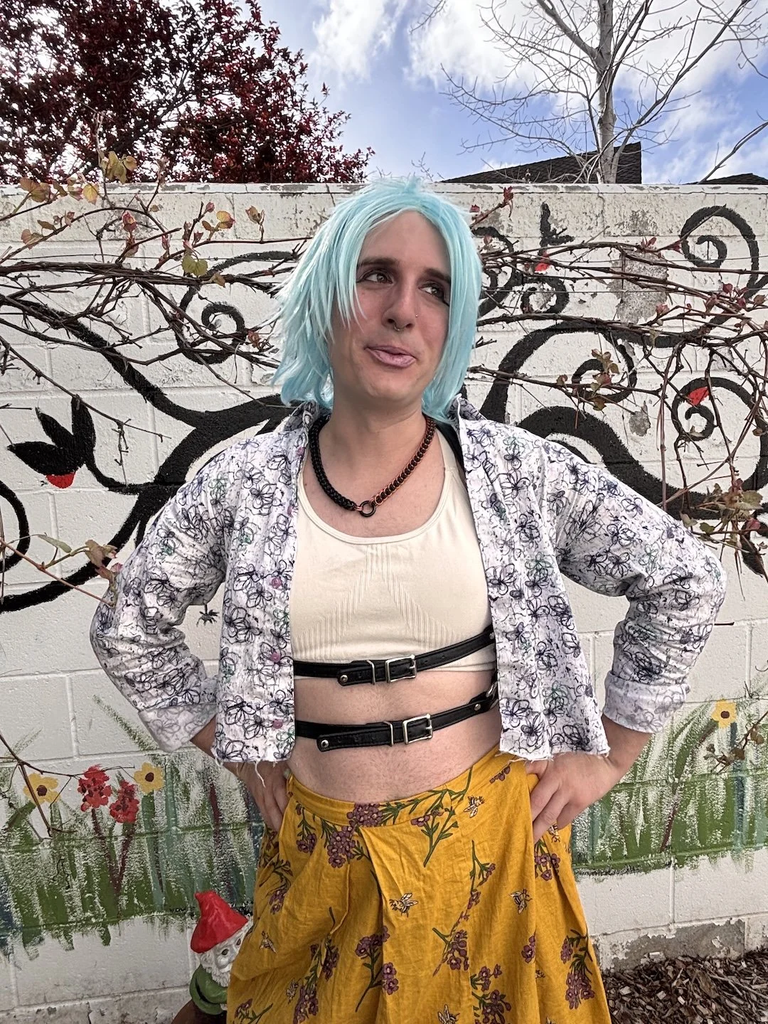 Transwoman with blue hair standing with her hands on her hips wearing an open button up, a leather harness, and long yellow skirt. She is standing against a white brick wall with a black tree painted on it