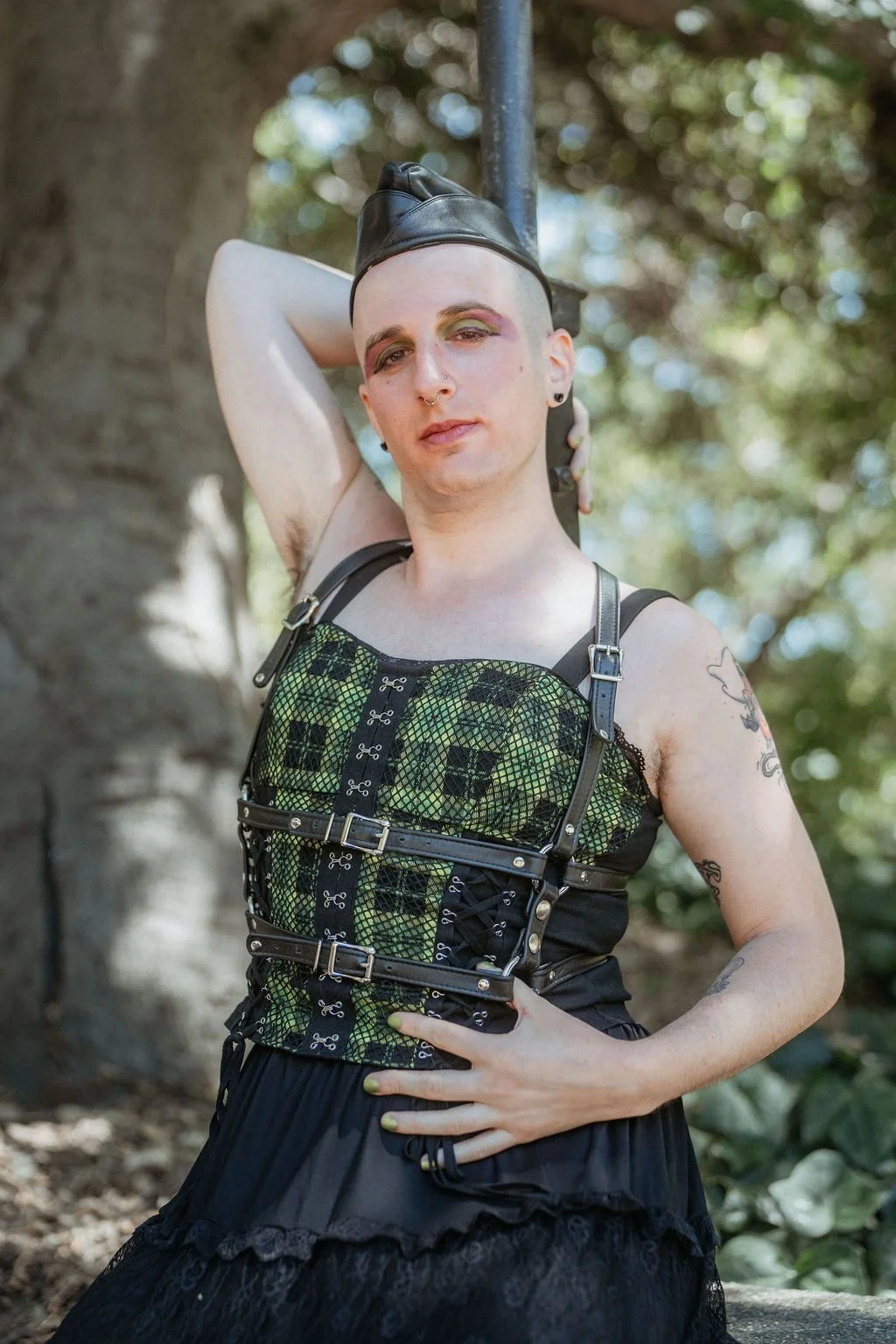 Calla wearing a leather cap, a black and green corset top with straps and buckles, standing outdoors with a tree and blurred foliage in the background.