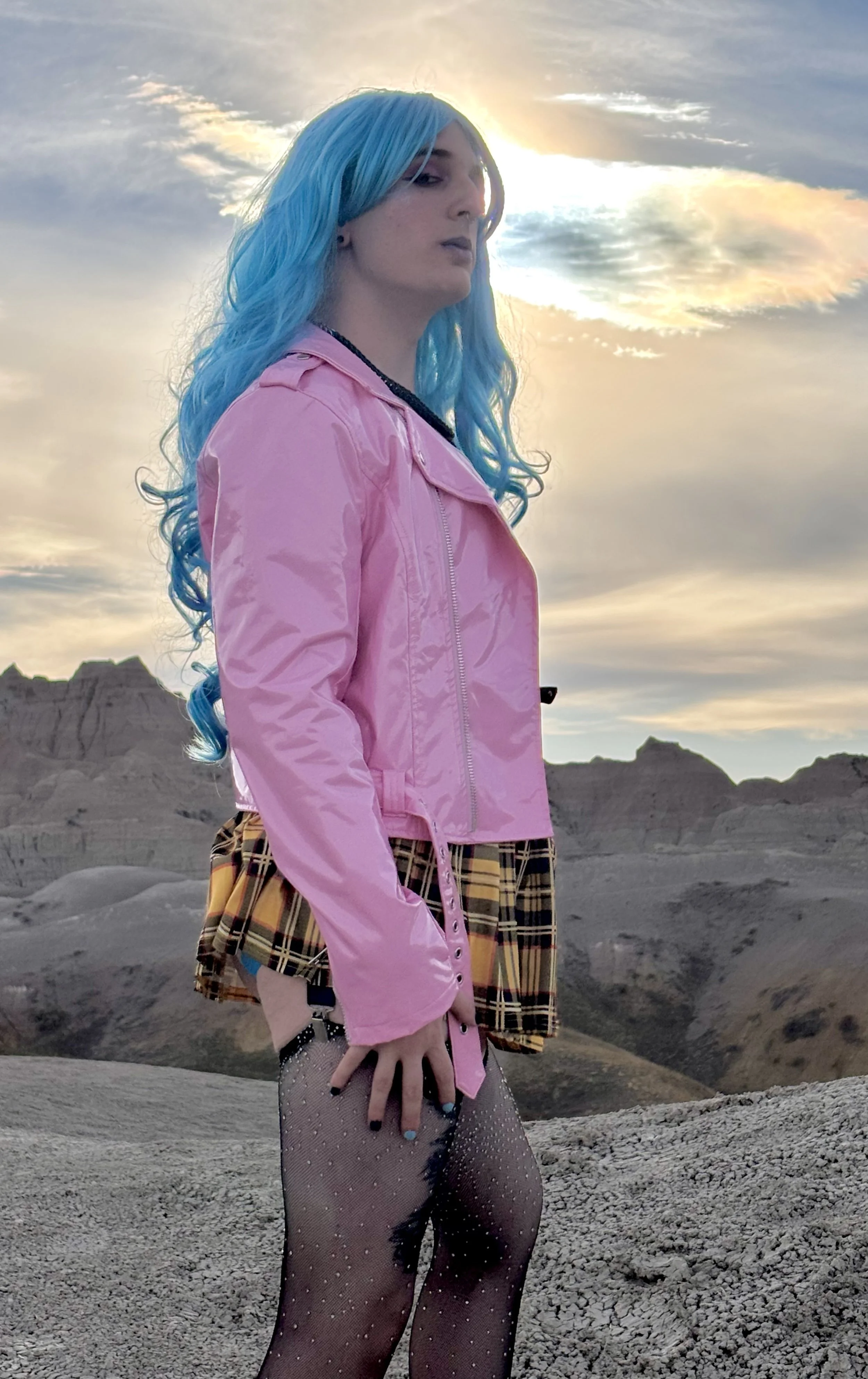 A woman with blue hair, a pink leather jacket, and a yellow plaid skirt posing powerfully at sunset in Badlands National Park in South Dakota
