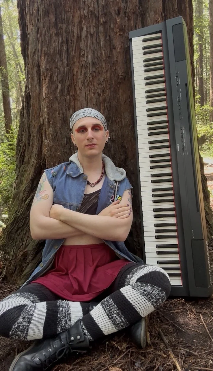 Transwoman sitting against a Redwood tree with her arms folded, her facial expression suggests she is waiting for you to show her something, her Yamaha keyboard is leaned against the tree next to her