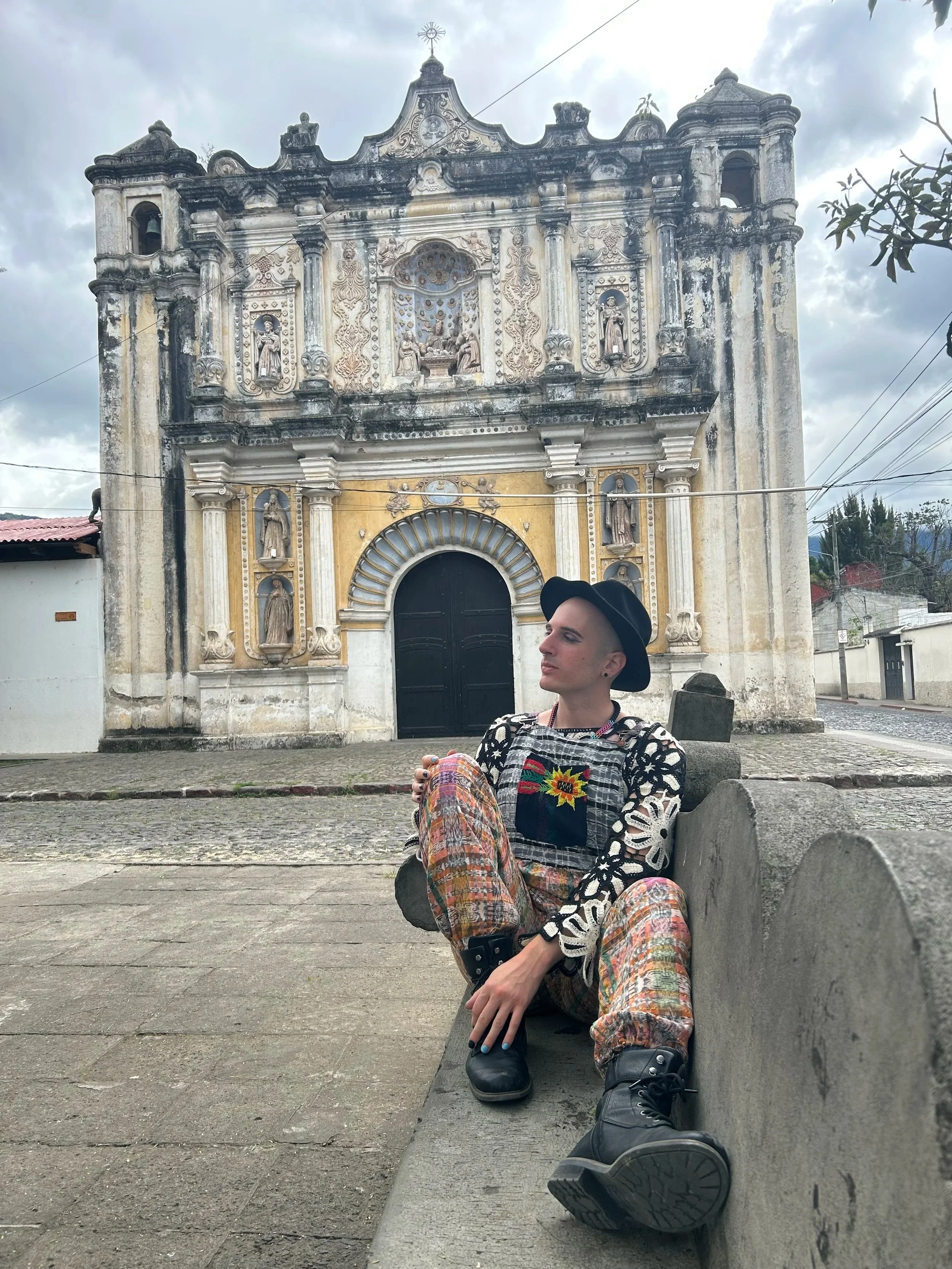 Person sitting on a bench in front of an old, ornate church building, wearing colorful patterned pants, a black wide-brimmed hat, and a black and embroidered top, with cloudy skies overhead.