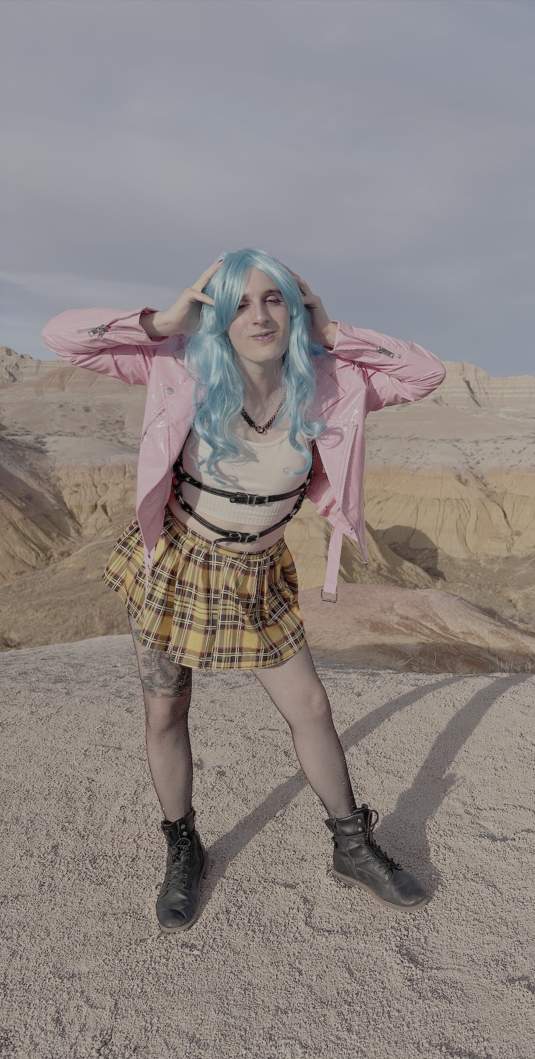 Trans woman with blue hair wearing a pink faux leather jacket, yellow plaid skirt, leggings, and boots with her hands on her head placed playfully, posing in Badlands National Park