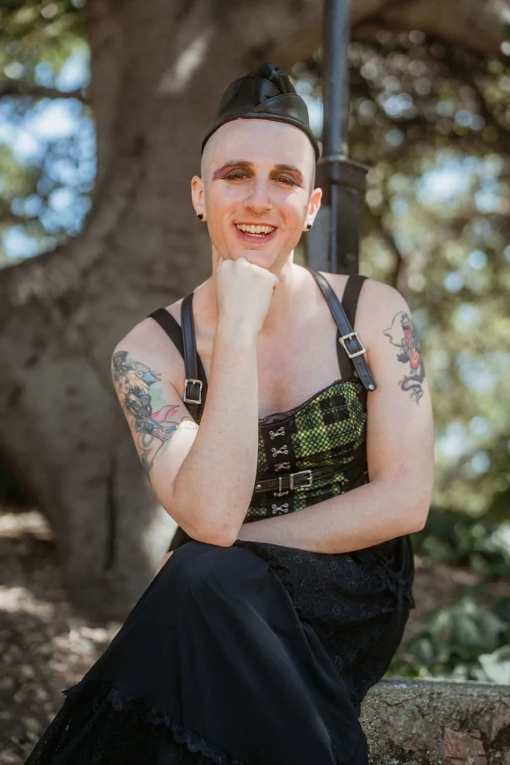 Calla, a reiki practitioner, smiling brightly while sitting on a cement block with her elbow resting on her knee and her chin resting on her fist. She's wearing a green plaid corset and a black skirt.blockstyle-border