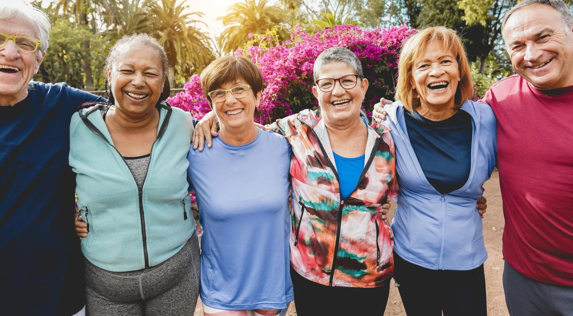 Six older adults standing outdoors with arms around each other, smiling, with pink flowers and palm trees in the background.