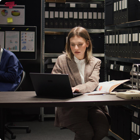A woman working on a laptop at a desk in an office or storage room surrounded by shelves with binders and folders.