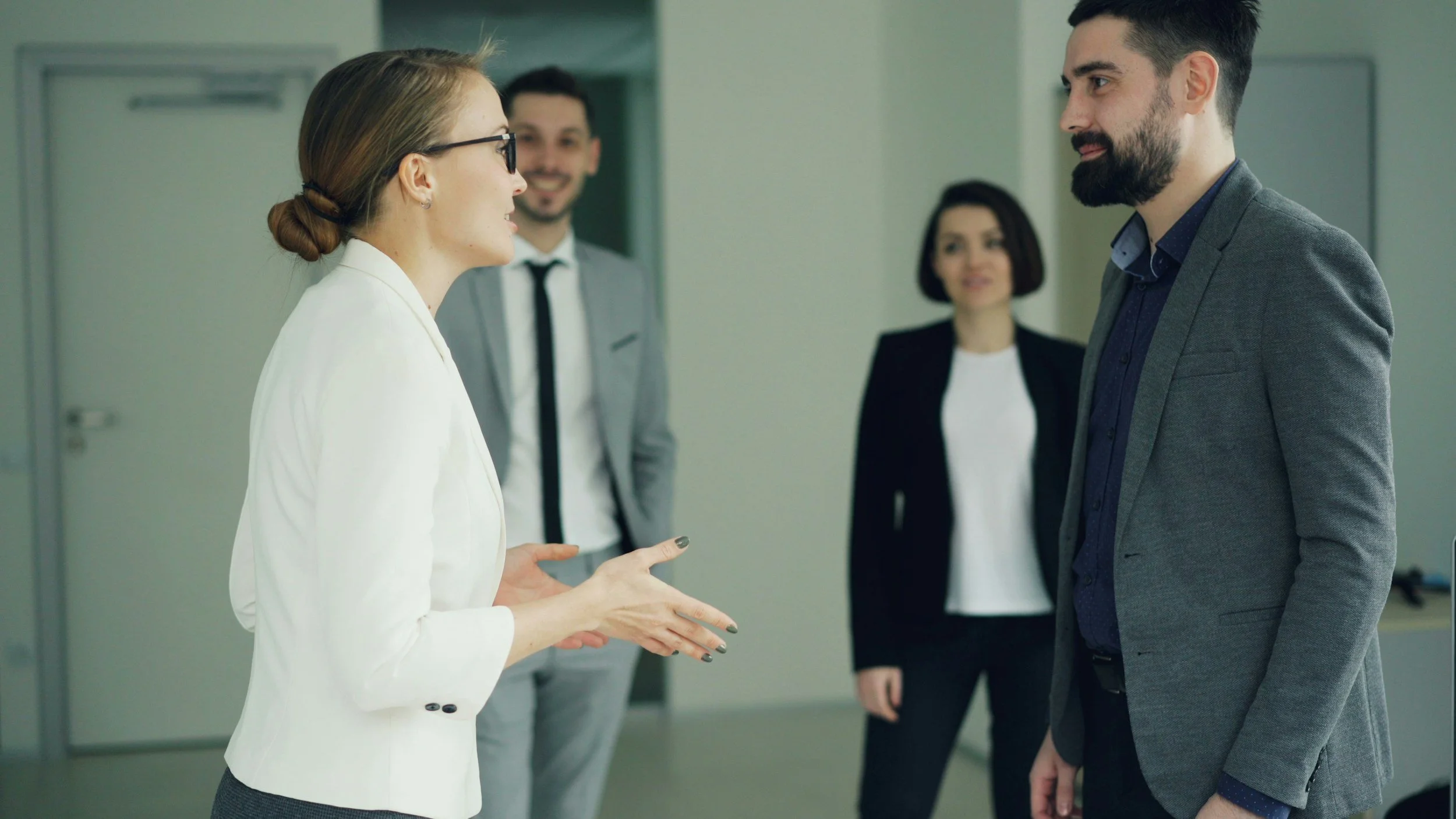 Four professionals are engaged in conversation in an office corridor. A woman with glasses and a white blazer is speaking to a bearded man in a grey suit. Two other professionals, a man in a grey suit and a woman with dark hair and a blazer, observe the interaction.