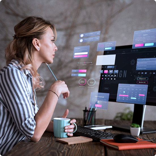 A woman in a striped shirt sitting at a desk interacts with holographic digital interface projections, with a computer monitor and keyboard, a mug, a notebook, and a plant nearby.