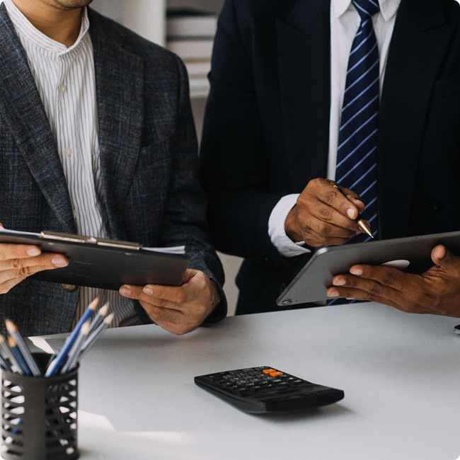 Two business professionals in suits working with digital devices at a desk, one holding a tablet and the other writing on a tablet, with a calculator and office supplies on the desk.