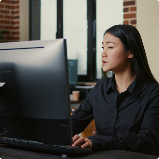 A woman with long black hair working on a computer in an office with brick and glass walls.