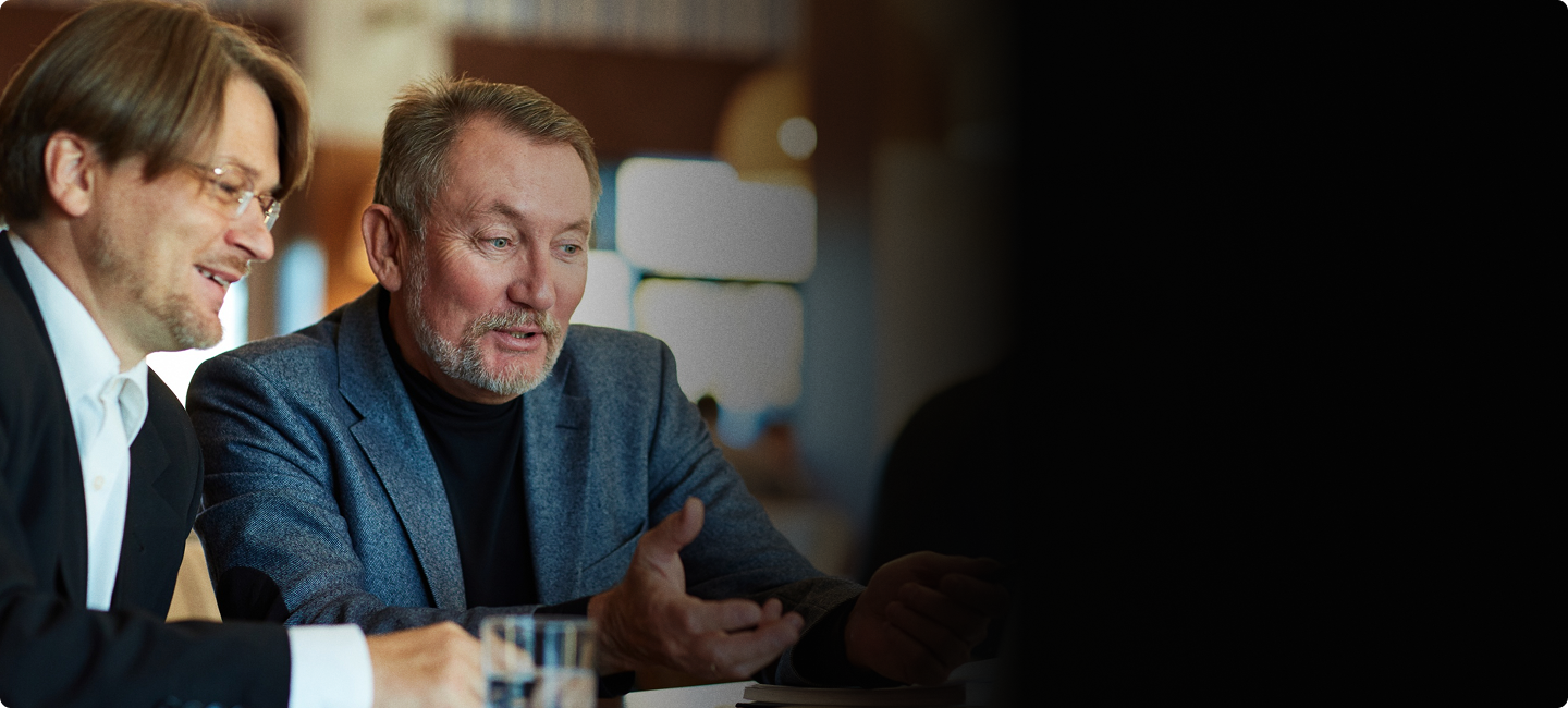 Two men are sitting at a table, engaged in a conversation. One man is talking while the other listens, with a glass of water in front of him. The setting appears to be a casual or professional environment.