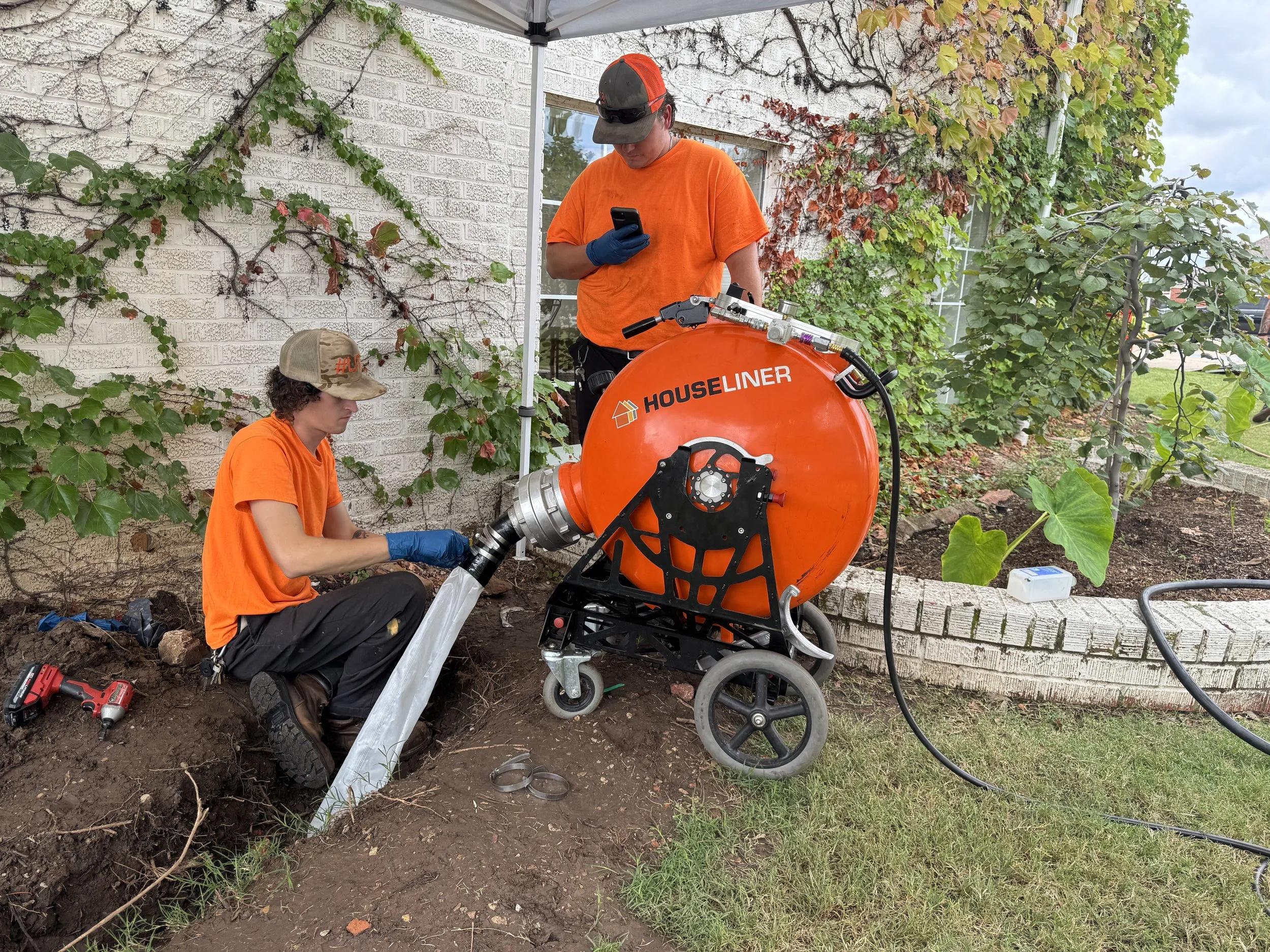 Two workers, one seated and one standing, working with a large orange machine labeled 'HOUSLINER' outdoors near a brick wall covered with green and red plants, installing or maintaining underground pipes, with tools and equipment nearby.
