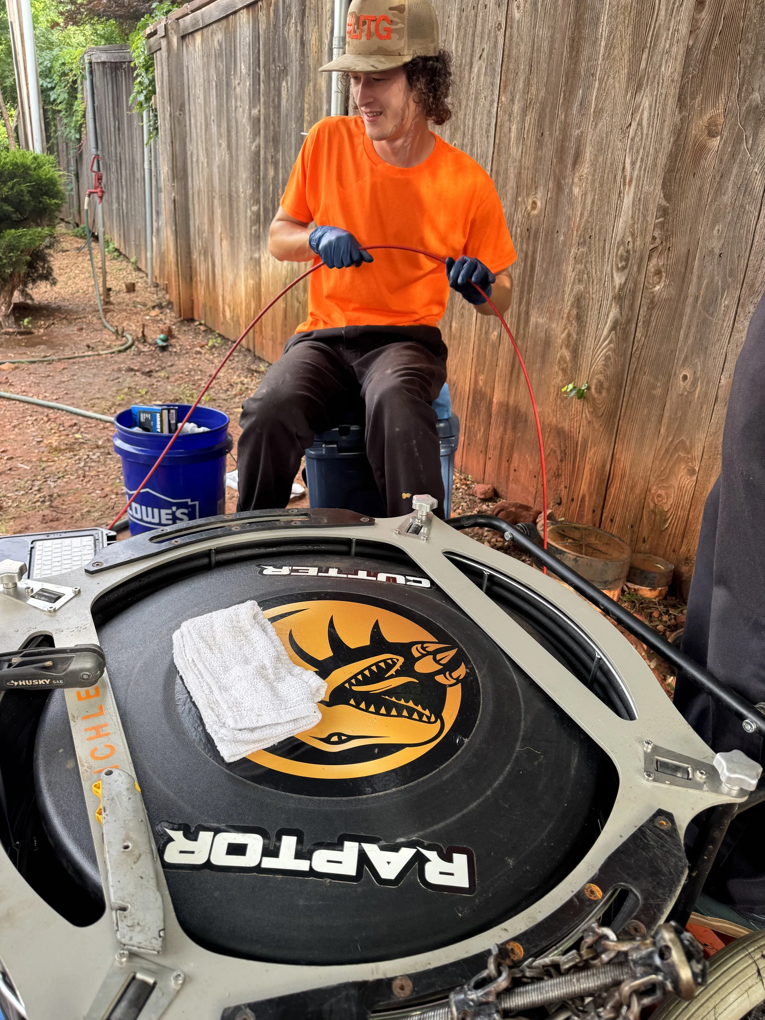 A person with curly hair wearing an orange T-shirt, black gloves, and a tan cap, sitting outdoors near a wooden fence, holding red wires, with a hockey puck and towel on a face-off circle with a mythical dragon logo.