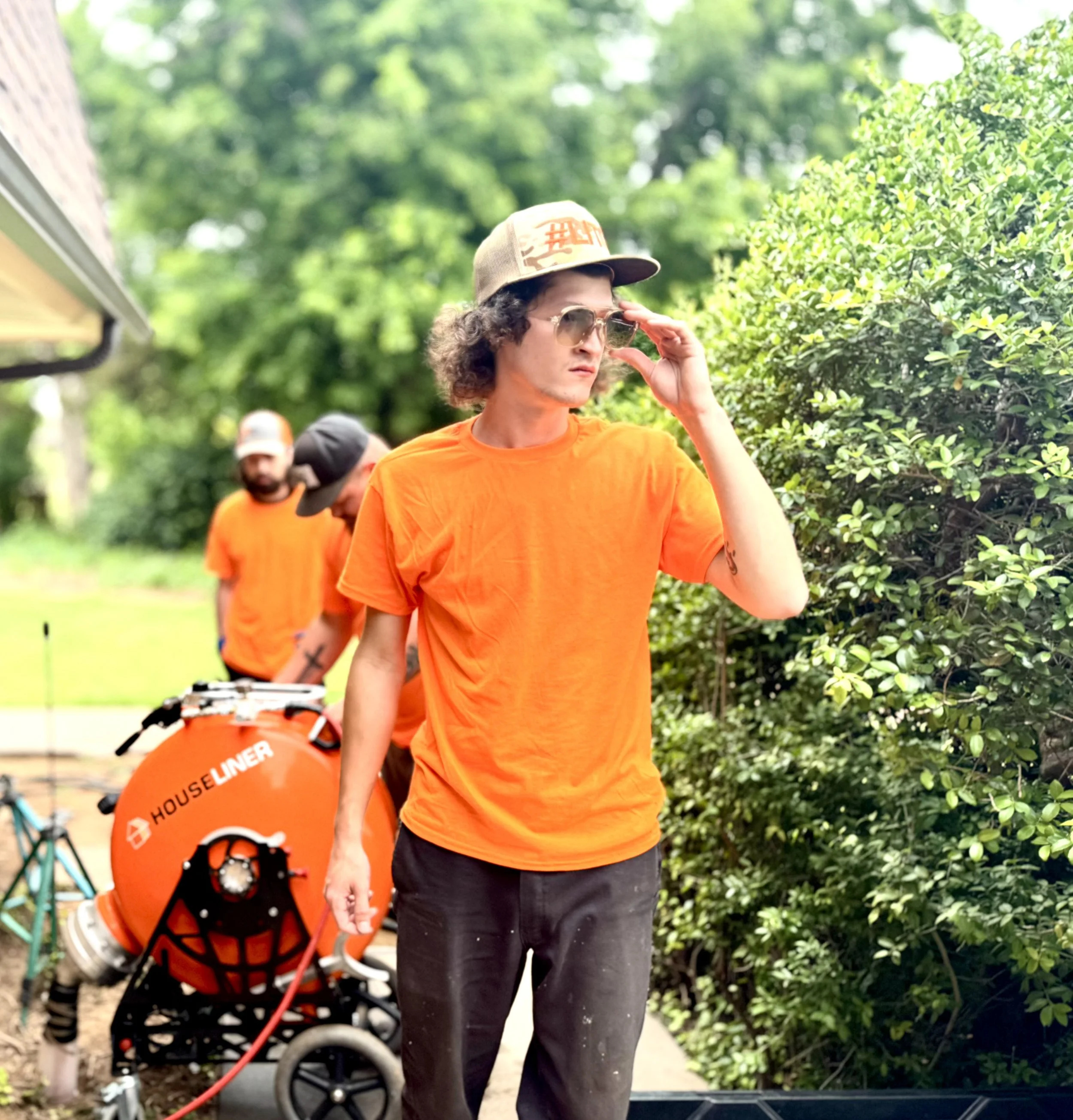 A young man with curly hair, sunglasses, and a cap adjusting his glasses with a bush on his right side, wearing an orange T-shirt. Two men in orange T-shirts and caps are in the background, one handling equipment near a hose reel labeled 'HOUSE LINER,' with greenery around.