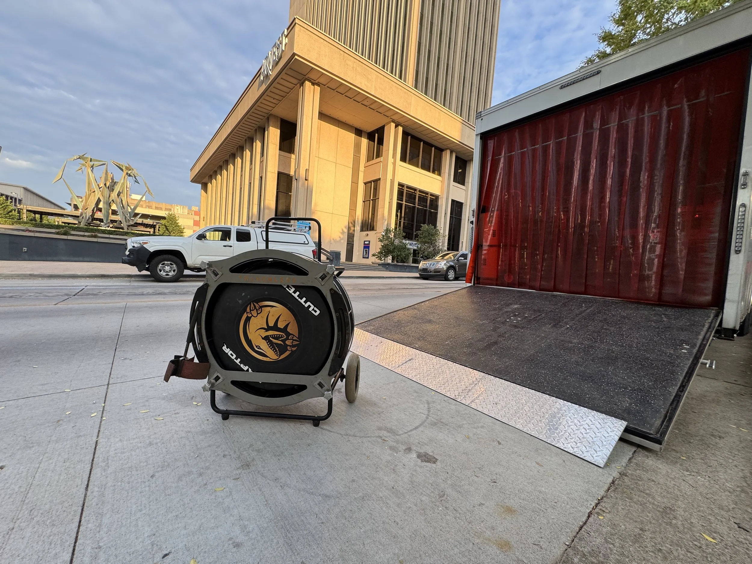 A delivery truck with its rear ramp lowered on a city sidewalk, a retractable hose reel with a dragon logo, and a building with classical architecture in the background.