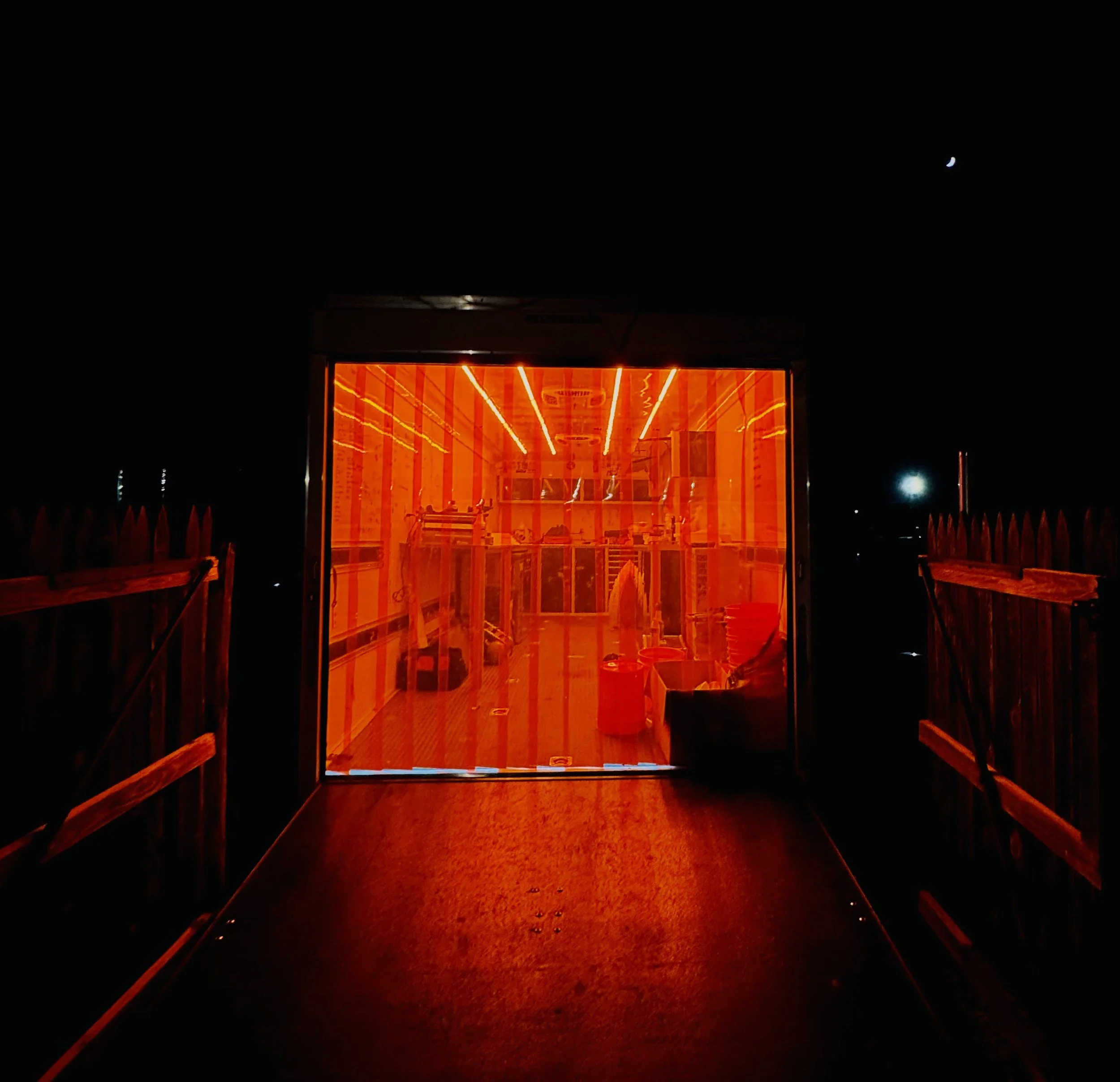 A doorway with a bright orange-lit interior, leading to a room filled with various objects, seen at night with a dark sky and a small crescent moon above.