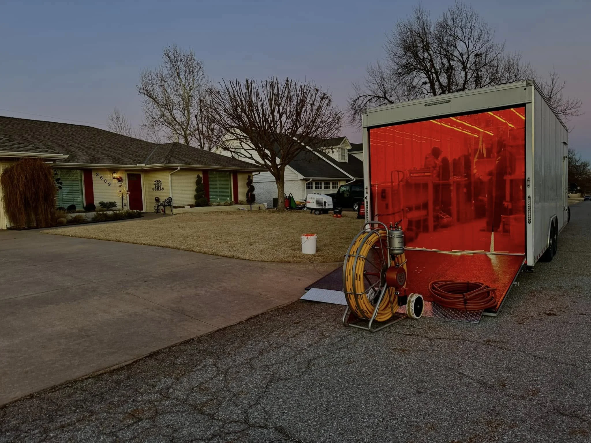 A mobile indoor construction or welding booth with a red interior, connected to yellow hoses and tools, parked on a residential street with houses and leafless trees, during dusk.