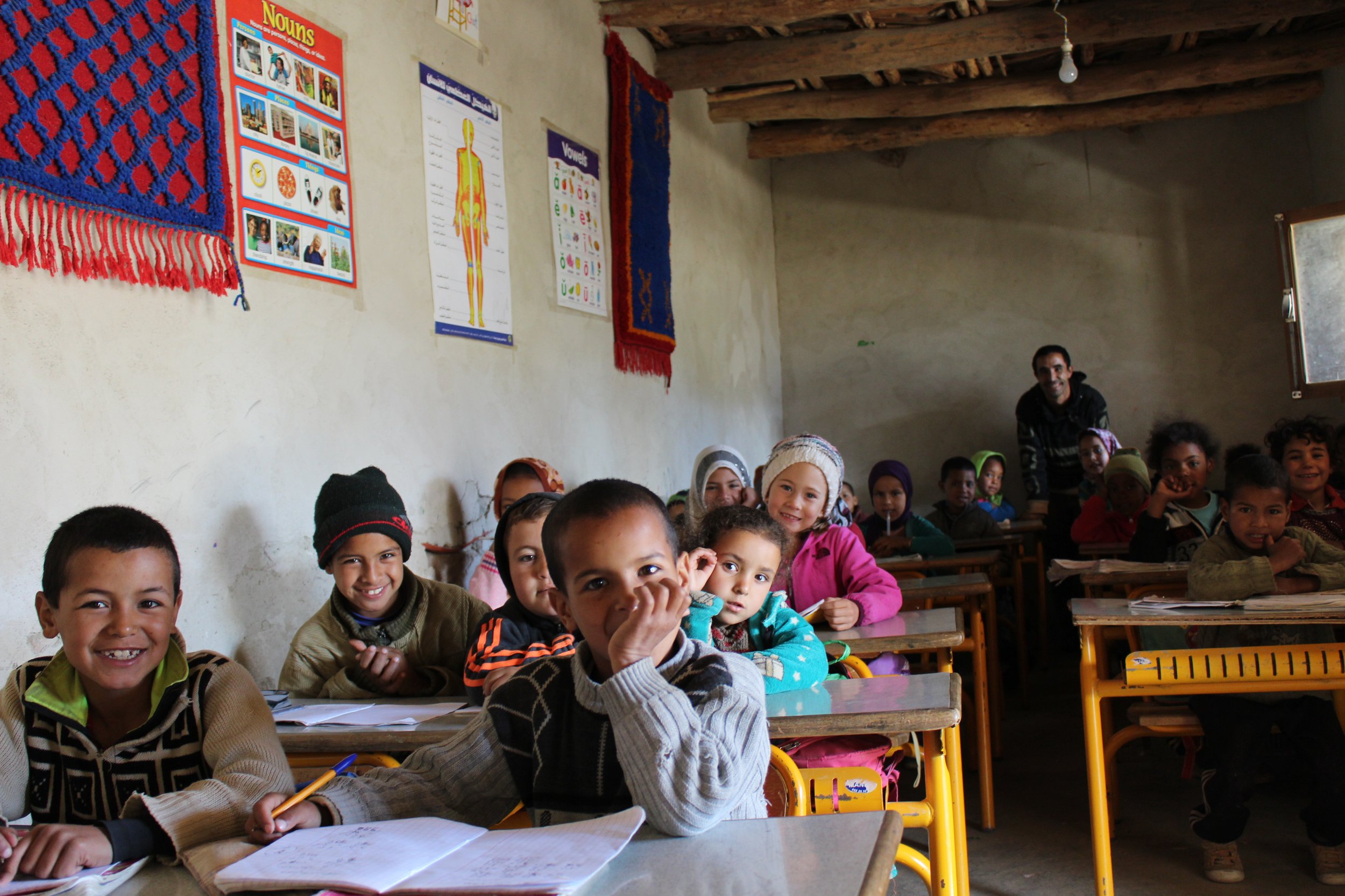 A classroom filled with young students sitting at desks, smiling, with educational posters on the wall.