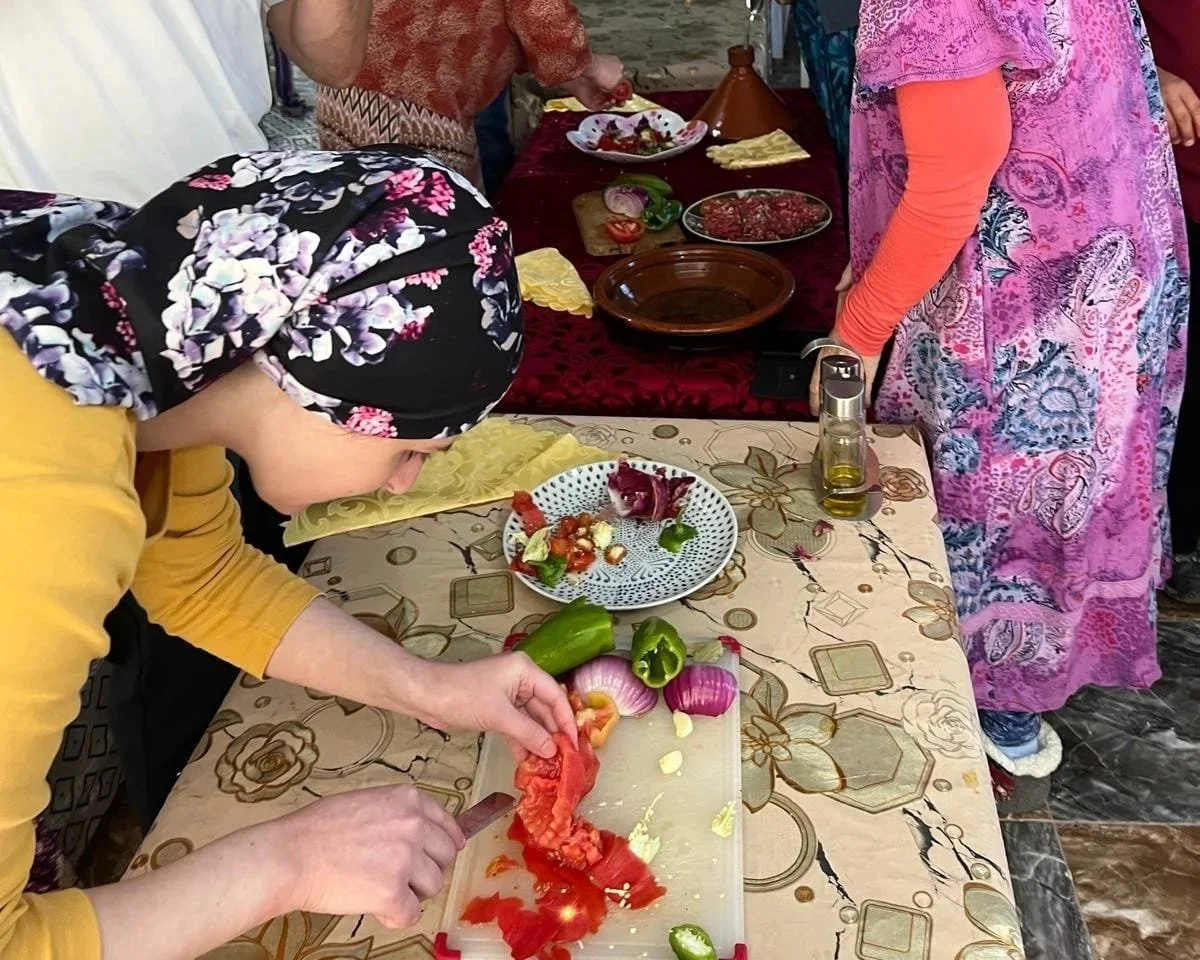 People preparing food on a table with vegetables such as tomatoes, onions, and peppers, and various plates and utensils.