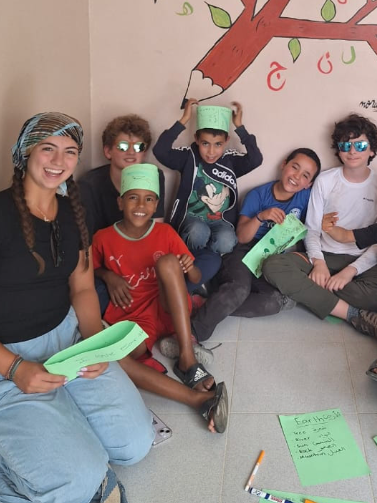 Group of kids and a woman sitting on the floor, smiling, with green paper hats and sheets, against a wall with a painted tree and writing.