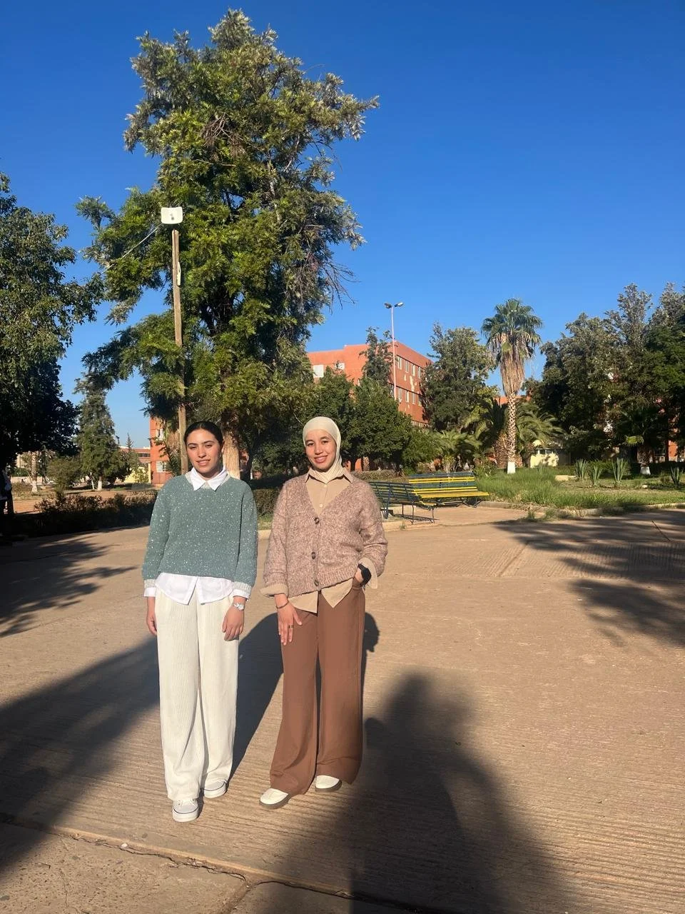 Two women standing outdoors on a sunny day with trees and buildings in the background.