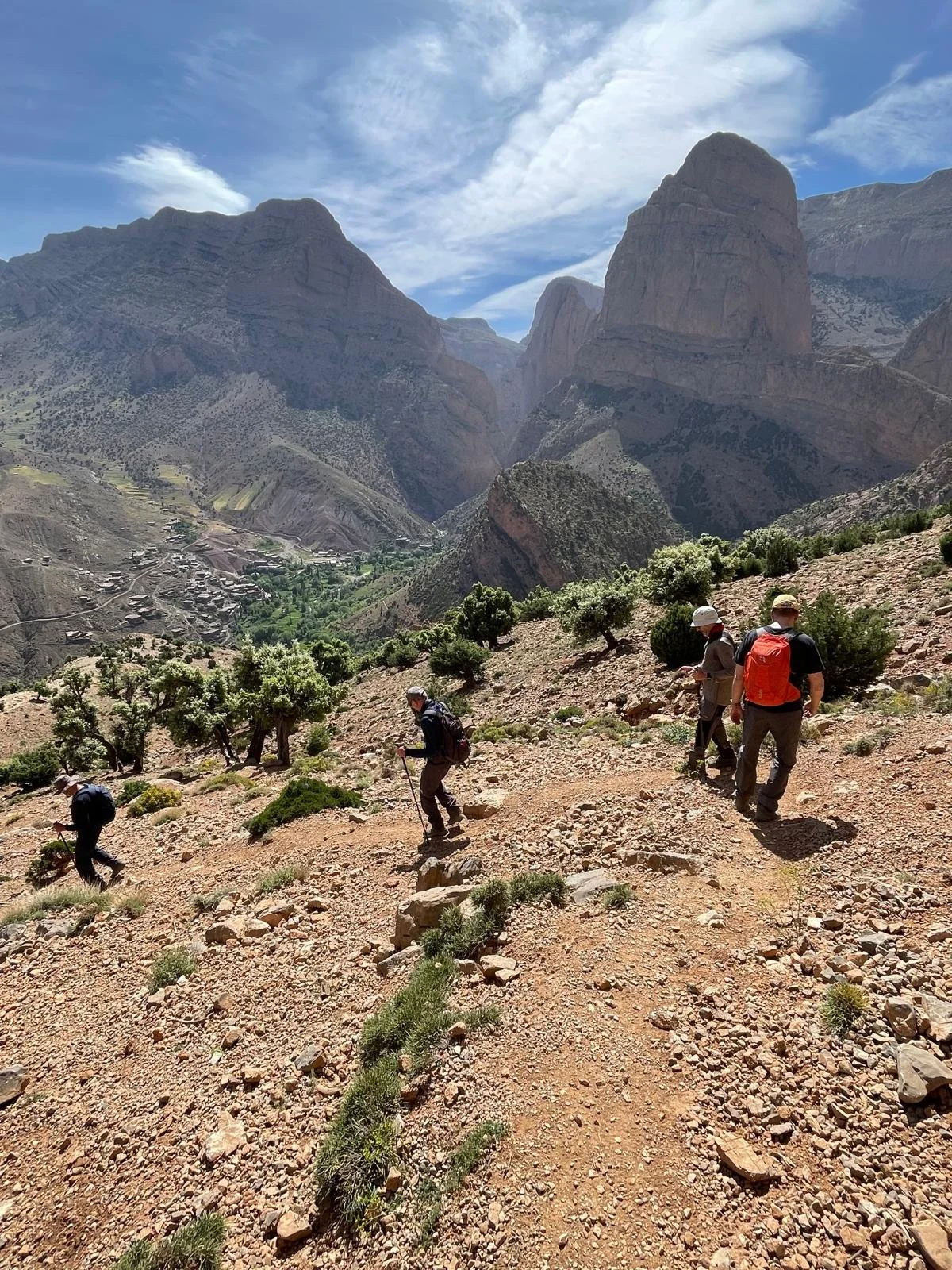 Hikers trekking along a rocky mountain trail in a valley surrounded by towering cliffs under a partly cloudy sky.
