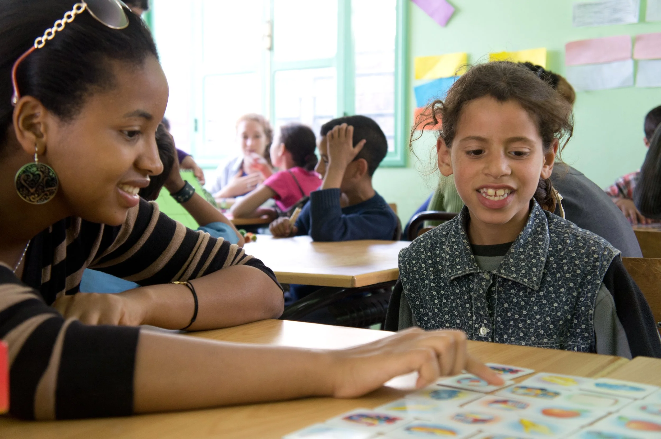 Two young girls are sitting at desks in a classroom, looking at each other and smiling. One girl has curly hair and a patterned shirt, the other has dark hair and large earrings. Other children are visible in the background, some with hands on their heads or talking. The classroom walls are painted green and decorated with colorful paper, and large windows let in natural light.