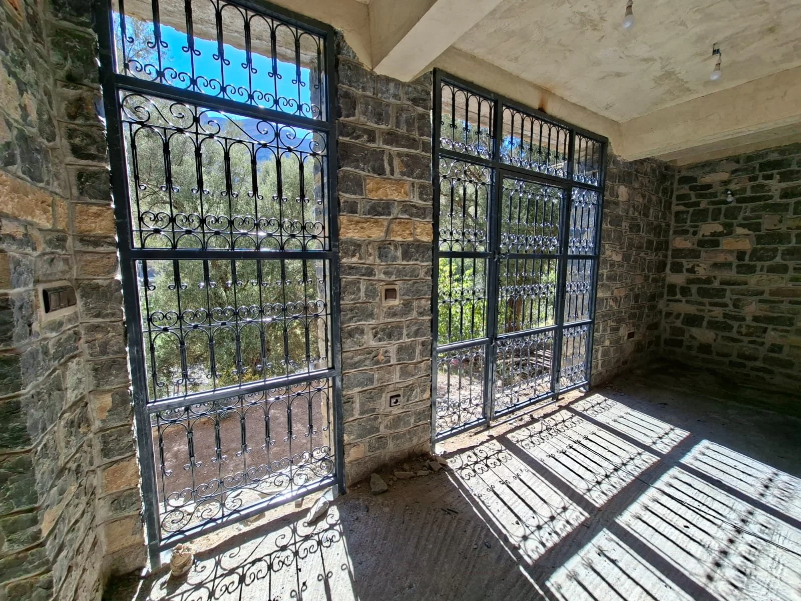 Interior of a building under construction with large windows and decorative black wrought iron security bars. Sunlight casts shadows on the unfinished concrete floor.