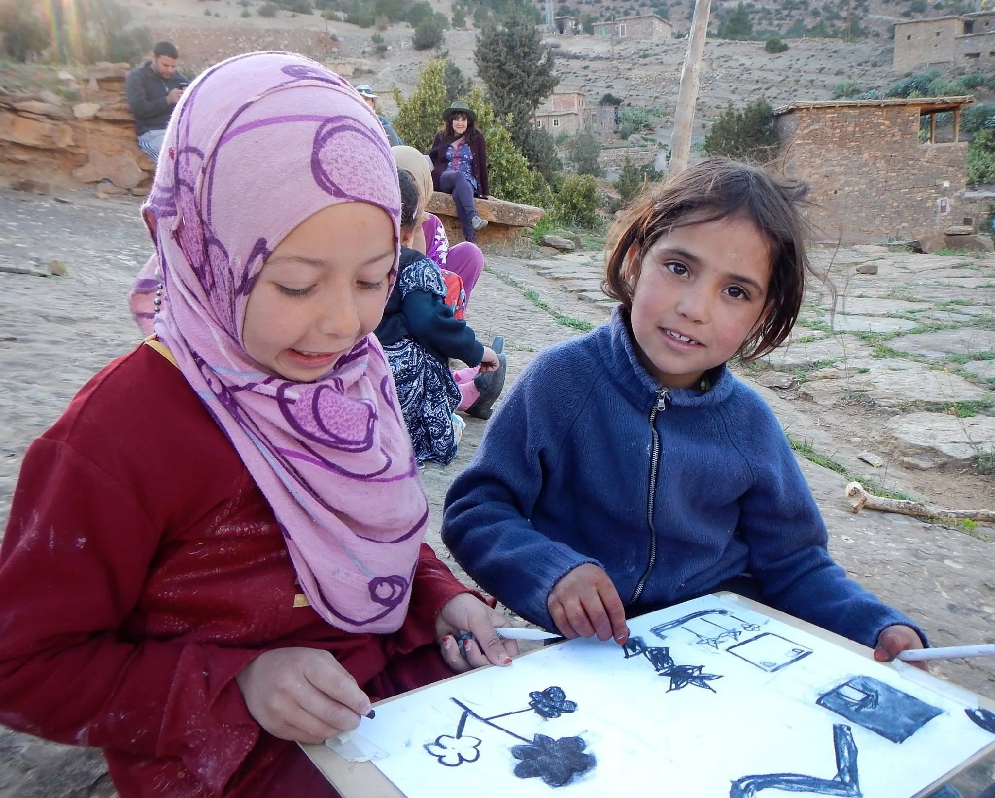 Children and a woman sitting outdoors, looking at drawings and sketches, with a woman sitting on a rock in the background. The scene is in a rural area with houses on a hillside.