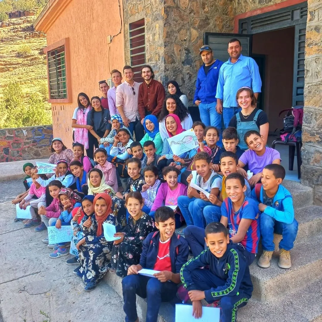 A group of children and adults smiling and sitting on steps outside a stone house, some children holding drawings, in a rural setting with green trees and mountains in the background.