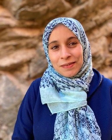 A woman wearing a blue patterned hijab and jacket, standing outdoors with a rocky background.