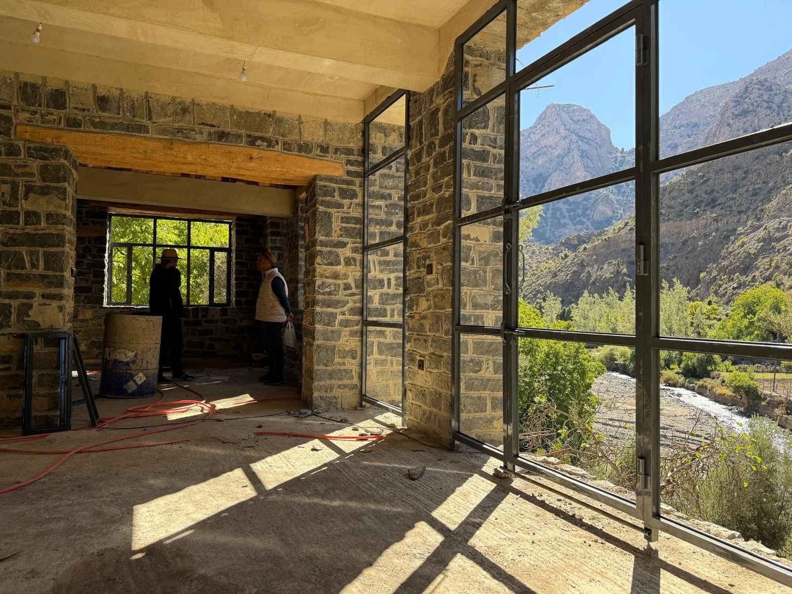 Interior of a building under construction with large windows overlooking mountains and a river. Two people are working inside, one pointing and the other holding a bag.